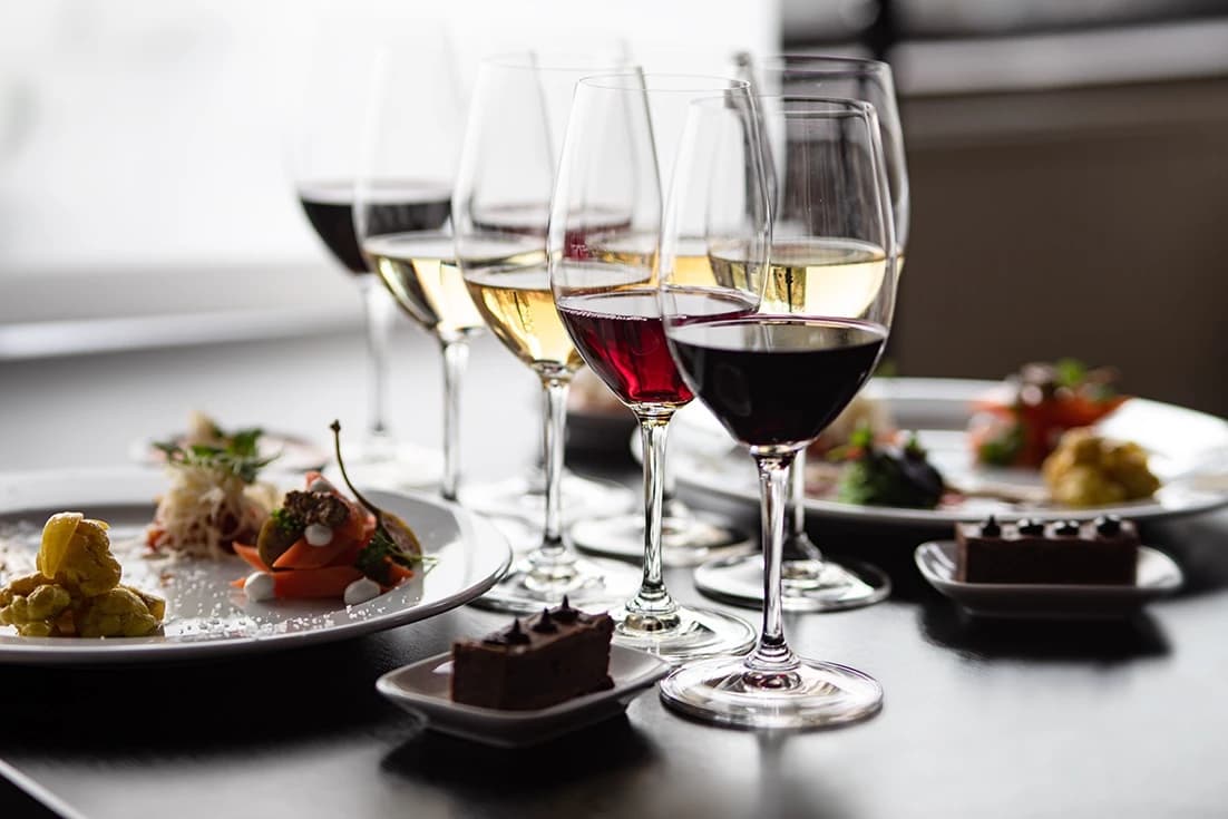 Flera vinglas med rött och vitt vin uppställda på ett bord, tillsammans med tallrikar och små rätter för en vinprovning.
